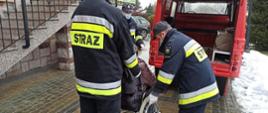 Trzech strażaków pomaga osobie poruszającej się na wózku inwalidzkim zająć miejsce w pojeździe