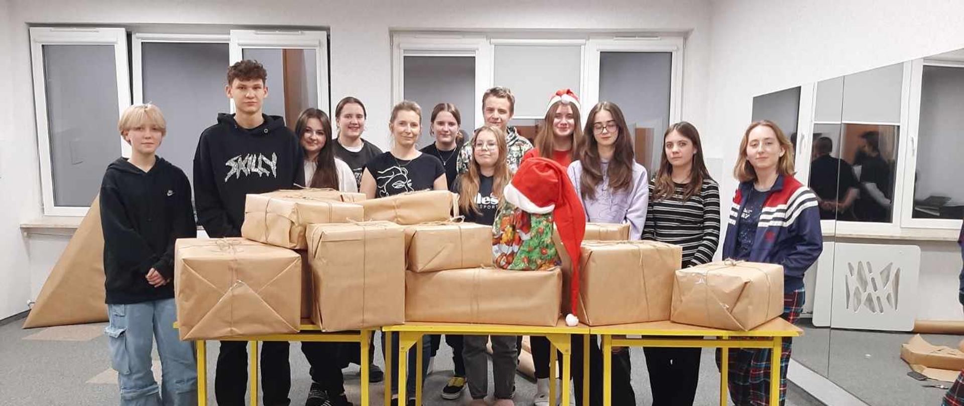 Kilkunastoosobowa grupa wychowanków z wychowawczynią stoi wokół leżących na stolikach zapakowanych paczek w świetlicy głównej, na środku paczek jest umieszczona duża czerwono-biała mikołajkowa czapka