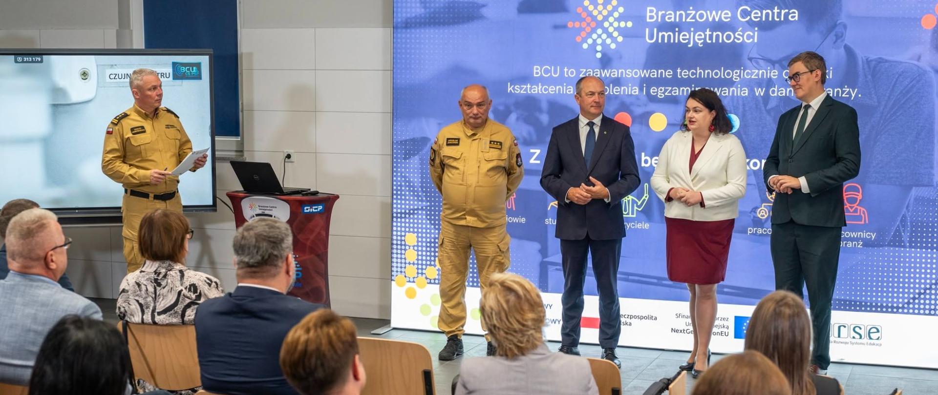 Czterech prelegentów — dwóch w mundurach i dwóch w strojach biznesowych — stoi przed dużym niebieskim ekranem z prezentacją o Branżowych Centrach Umiejętności, przemawiając do siedzących uczestników.