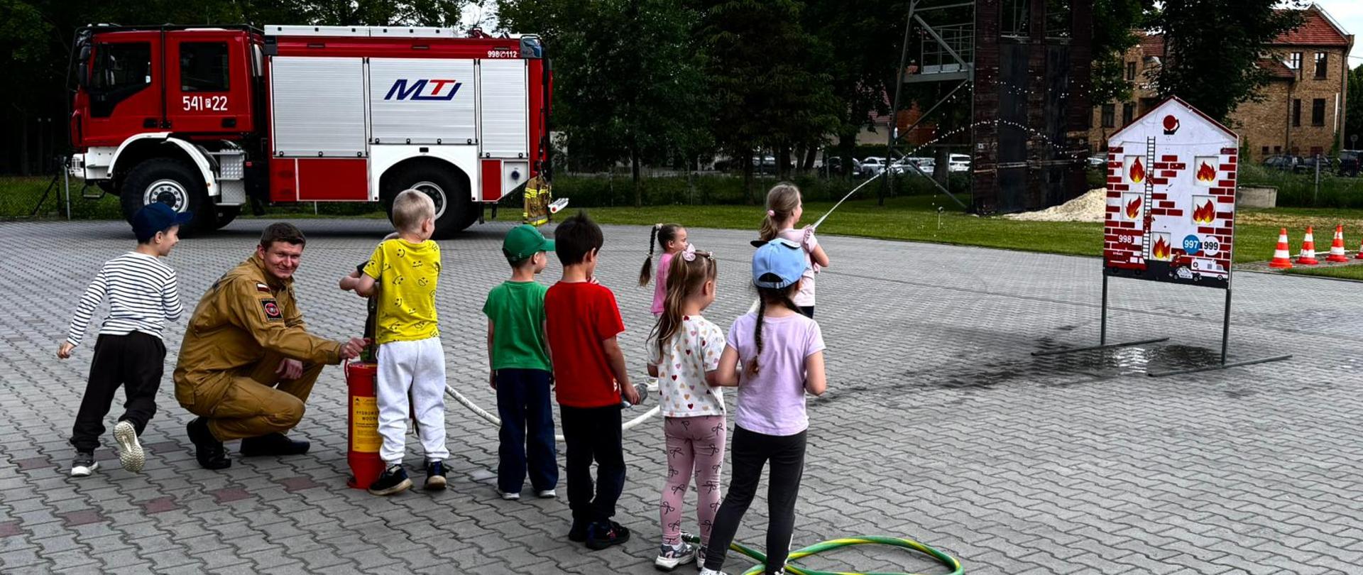 Dzieci w trakcie konkurencji na placu manewrowym. W tle wóz strażacki i zadrzewienie.