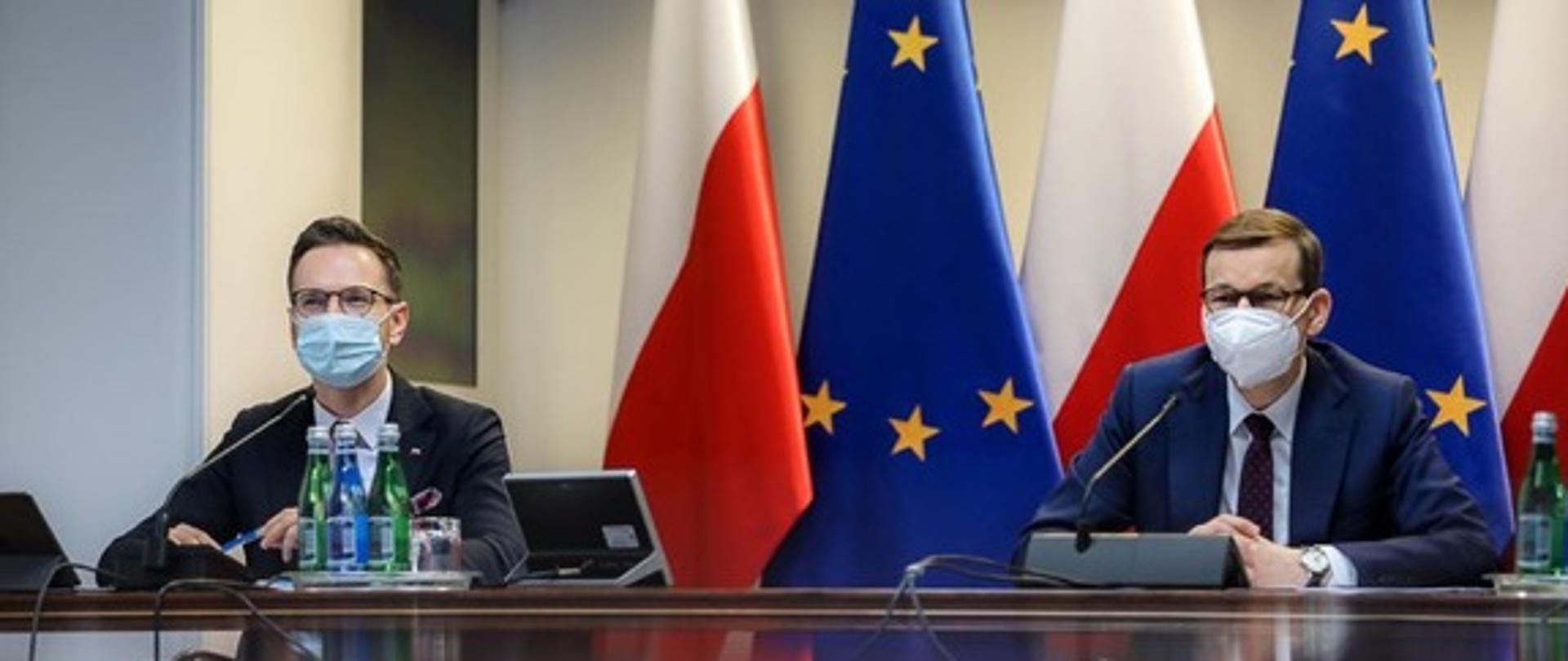 Przy stole konferencyjnym siedzą wiceminister Waldemar Buda oraz premier Mateusz Morawiecki. Za ich plecami flagi Polski i UE