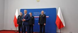 Na pierwszym planie wojewoda kujawsko-pomorski, za nim strażacy w mundurach wyjściowych - dwóch PSP, jeden OSP. Za plecami ścianka medialna z logo Urzędu Wojewódzkiego.