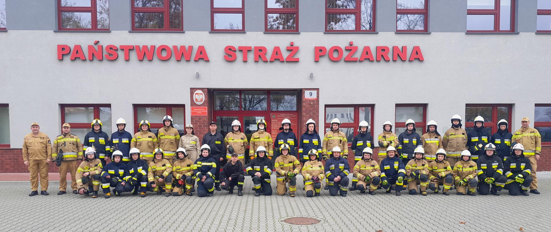 Grupa strażaków w dwuszeregu - pierwszy szereg kuca, drugi szereg stoi. z tyłu budynek z napisem Państwowa Straż Pożarna.