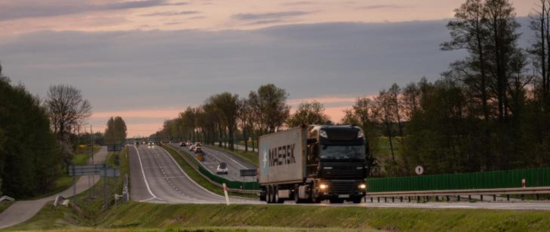 Droga ekspresowa o dwóch pasach dla każdego kierunku. Na pierwszym planie widoczna ciężarówka przewożąca kontener. W tle inne pojazdy, w tym także samochody osobowe.