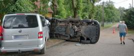 Na pierwszym planie widoczne są dwa samochody typu Minibus. Jeden z nich leży na boku, natomiast drugi jest wbity w leżący samochód. W tle widać drzewa oraz sieć linii energetycznych. Droga gruntowa zabezpieczona jest pachołkiem ostrzegawczym, po prawej stronie mężczyzna. 