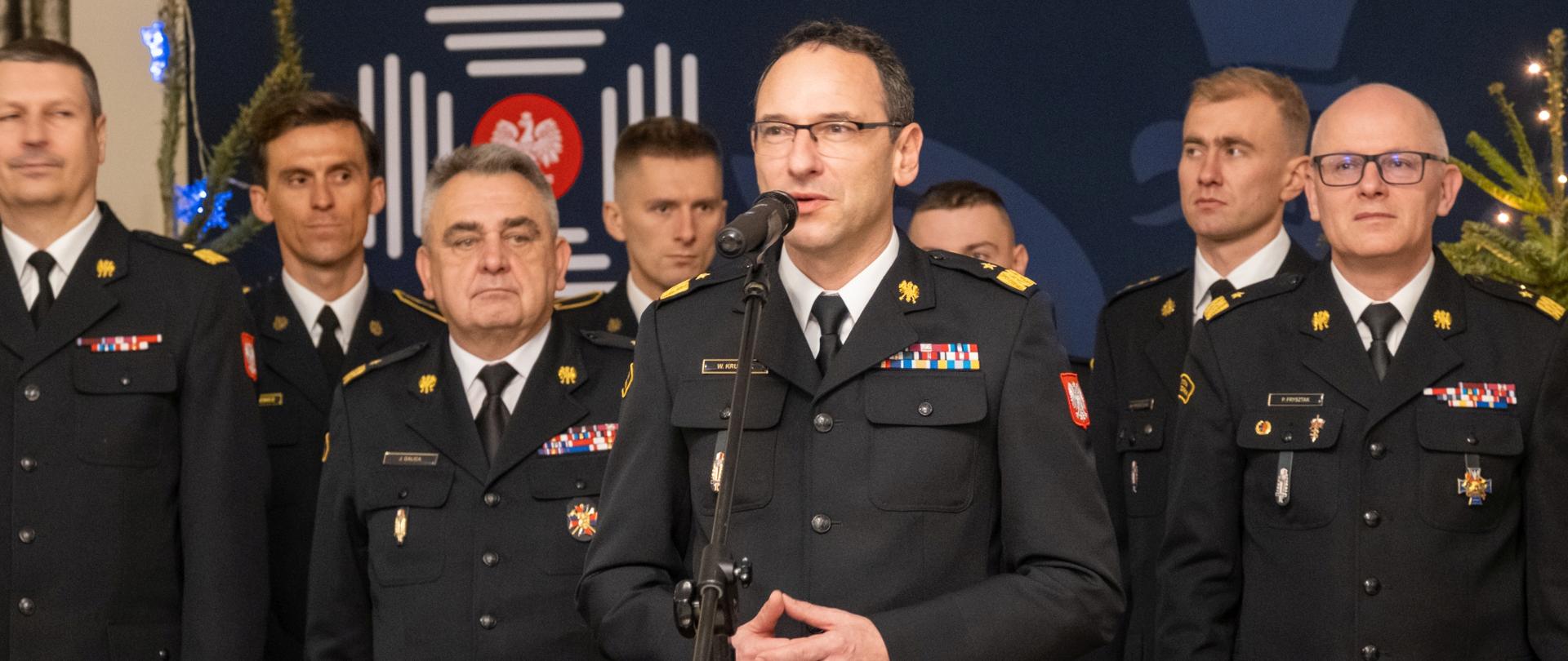Przemawiający oficer PSP w galowym mundurze, otoczony przez grupę strażaków, w tle świąteczne dekoracje i godło Państwowej Straży Pożarnej.
