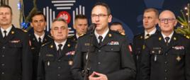 Przemawiający oficer PSP w galowym mundurze, otoczony przez grupę strażaków, w tle świąteczne dekoracje i godło Państwowej Straży Pożarnej.