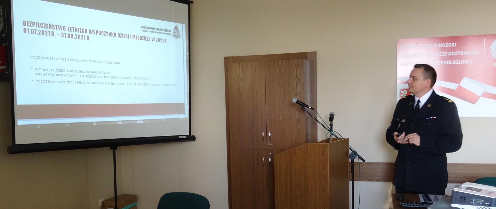 Zastępca Komendanta Powiatowego PSP stoi przed ekranem projektora, przedstawiając informacje na temat bezpieczeństwa letniego podczas wypoczynków dzieci i młodzieży. 