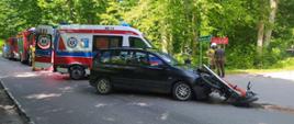 Na asfalcie leży czerwony motocykl, który zderzył się z szarym pojazdem osobowym. Obok stoją policjanci oraz karetka pogotowia.