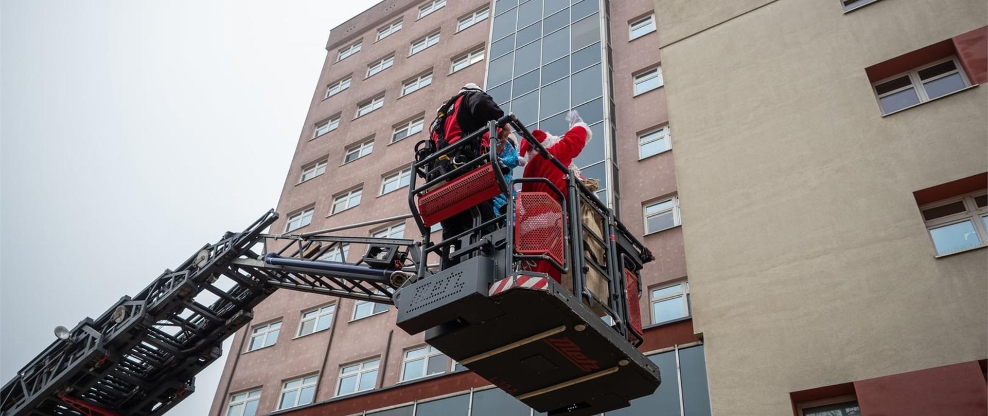 Na pierwszym planie kosz drabiny mechanicznej, w koszu święty mikołaj i strażak, w tle budynek szpitala i zjeżdżający po elewacji budynku na linie alpinistycznej ratownik wysokościowy