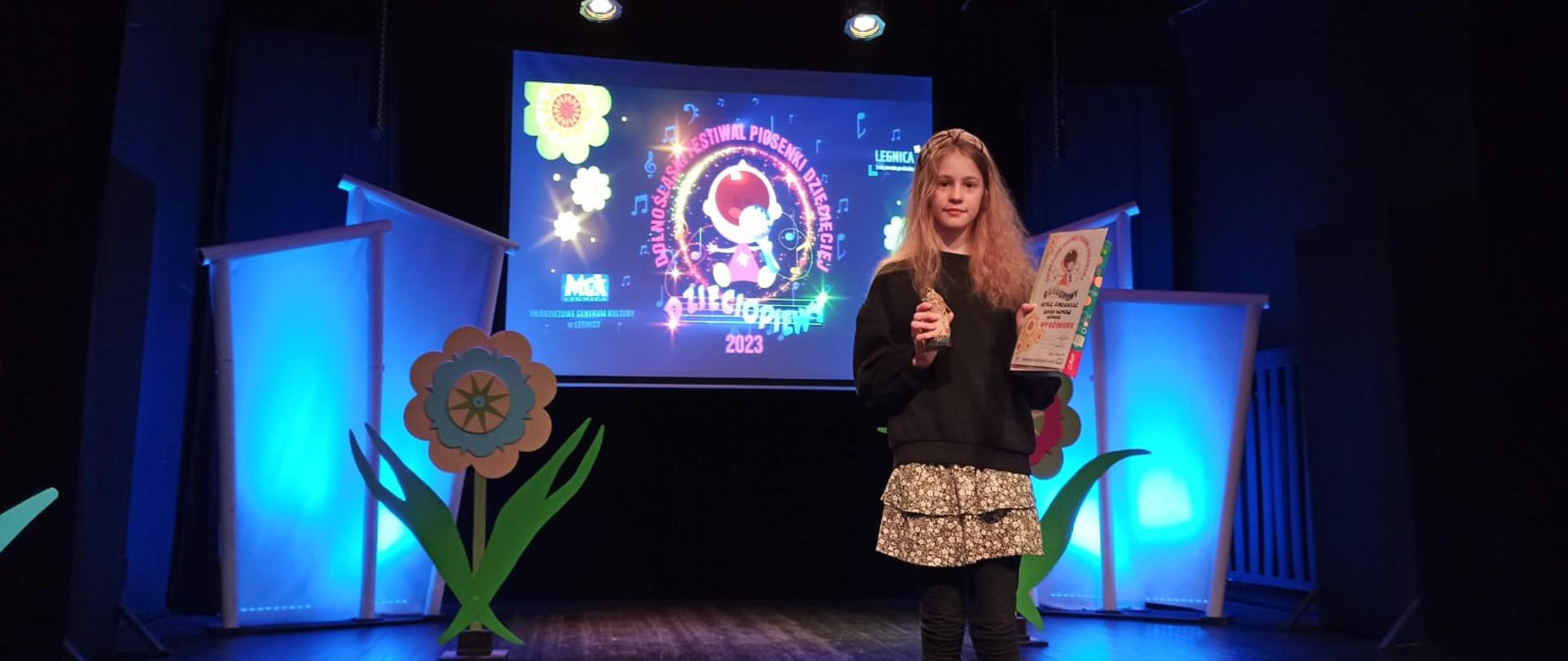 Zdjęcie przedstawia dziewczynę stojąca na scenie, trzymającą w rękach dyplom z konkursu i statuetkę. W tle ekran wyświetlający nazwę konkursu i elementy dekoracyjne.