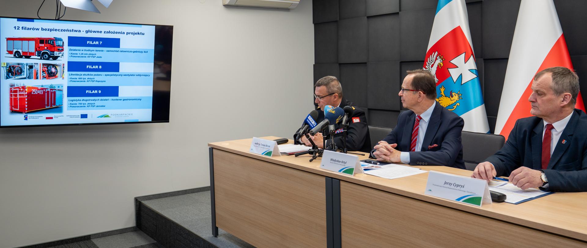 Konferencja prasowa w Urzędzie Marszałkowskim Województwa Podkarpackiego dotycząca nowej architektury bezpieczeństwa regionu.
Postacie i układ sceny
Na zdjęciu, przy jasnym drewnianym stole na podwyższeniu, siedzą trzej uczestnicy konferencji:
Z lewej: Nadbrygadier Tomasz Baran, Podkarpacki Komendant Wojewódzki PSP, ubrany w ciemny mundur służbowy.
W środku: Marszałek Władysław Ortyl w ciemnym garniturze i czerwonym krawacie.
Z prawej: Jerzy Cypryś, Przewodniczący Komisji Bezpieczeństwa Publicznego i Zatrudnienia, w ciemnej marynarce.
Przed prelegentami ustawione są mikrofony mediów oraz identyfikatory z nazwiskami.
Prezentacja projektu
Po lewej stronie kadru znajduje się duży monitor z wyświetloną planszą tytułową: „12 filarów bezpieczeństwa – główne założenia projektu”. Na ekranie widoczne są grafiki przedstawiające:
Filar 7: Samochód ratowniczo-gaśniczy 4x4 dla KP PSP Jasło.
Filar 8: Specjalistyczny wentylator oddymiający dla KP PSP Ropczyce.
Filar 9: Kontener gastronomiczny dla KP PSP Jarosław.
Tło i detale techniczne
Za uczestnikami znajdują się dwie flagi: województwa podkarpackiego oraz biało-czerwona flaga Polski.
Ściana w tle wykonana jest z ciemnych, dekoracyjnych paneli o strukturze 3D.
W lewym górnym rogu widoczna jest profesjonalna lampa studyjna typu softbox, doświetlająca stół.
W dolnym rogu zdjęcia widnieje nota autorska: „© Sebastian Stankiewicz | Biuro prasowe UMWP”.
