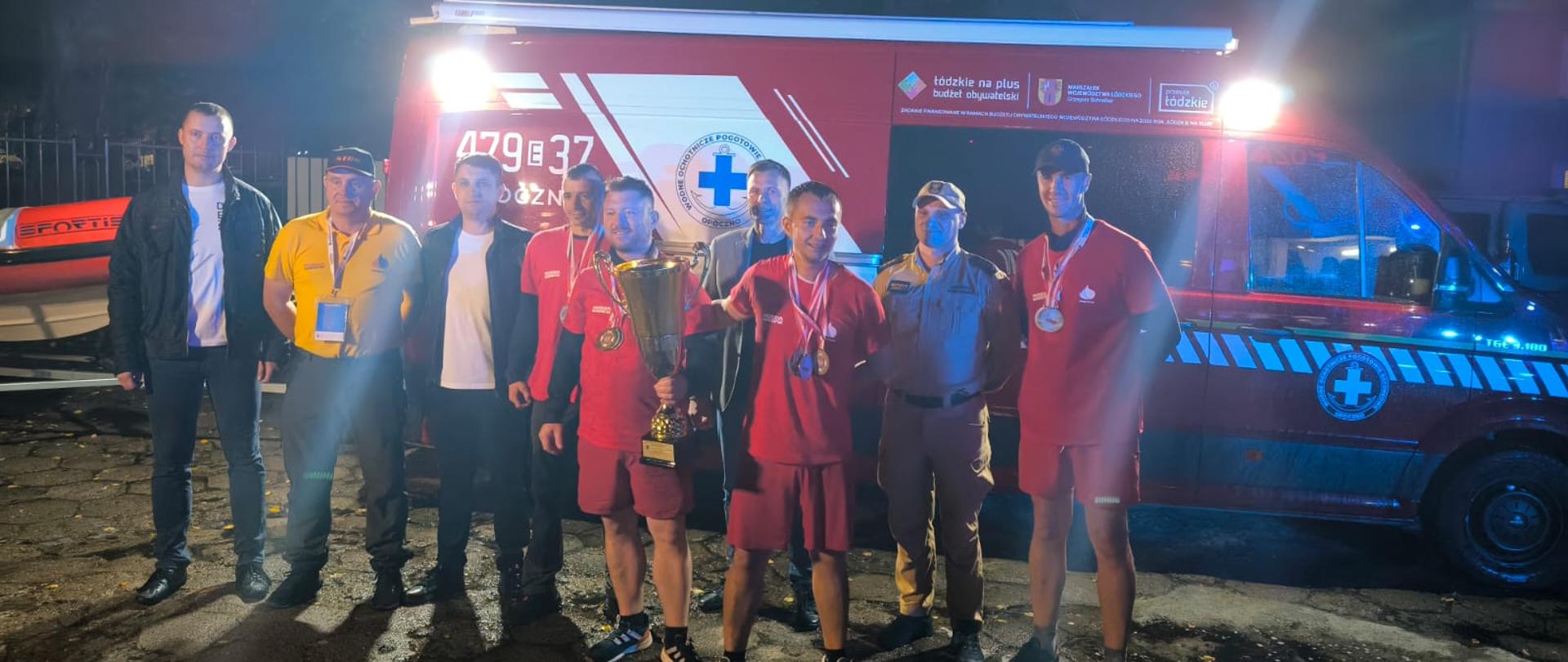 Pora nocna, czterech strażaków ochotniczej straży pożarnej ubranych w stroje sportowe koloru czerwonego, po środku trzymają puchar z mistrzowski, z założonymi medalami pozują szczęśliwi w towarzystwie komendanta powiatowego oraz innych przedstawicieli Państwowej Straży Pożarnej oraz urzędu miejskiego. W tle oświetlony samochód ratownictwa wodnego. 