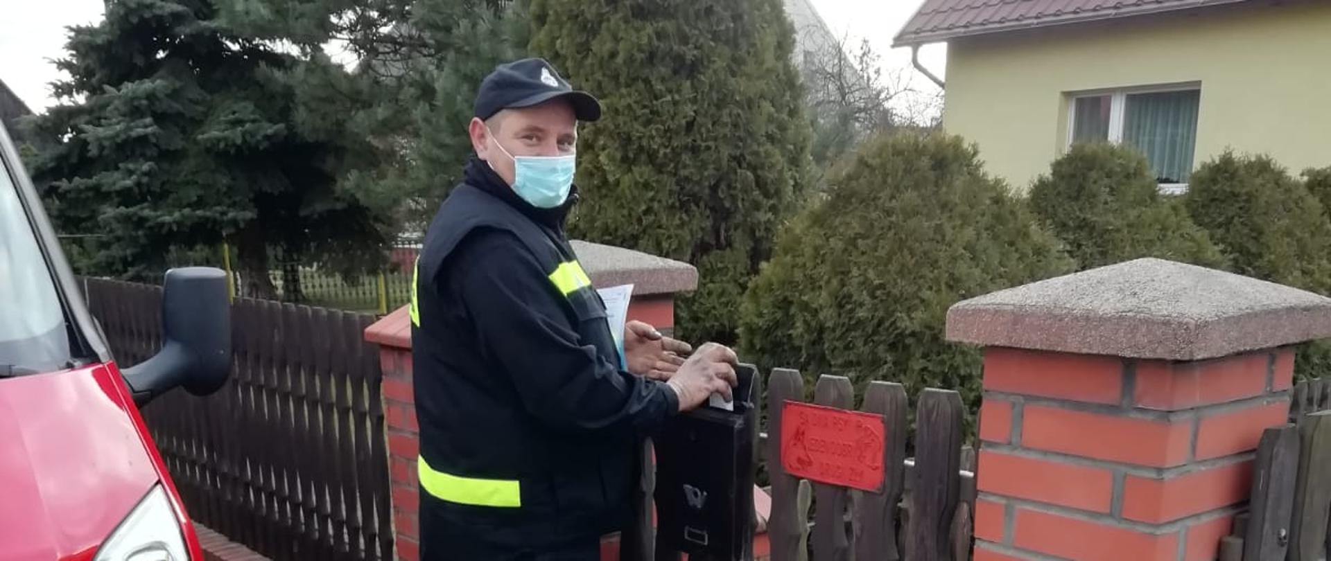 Na zdjęciu widać druha z jednostki Ochotniczej Straży Pożarnej w umundurowaniu koszarowym, stojącego przy posesji, który wrzuca ulotkę dotyczącą szczepień do skrzynki znajdującej się na ogrodzeniu. Z lewej strony widać samochód pożarniczy z jednostki Ochotniczej Straży Pożarnej. W tle widać dom i ogród.
