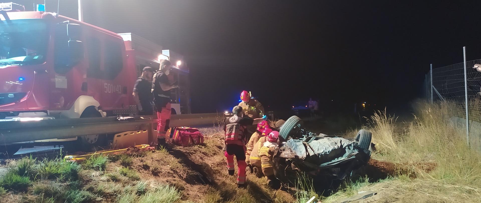 strażacy podczas ewakuacji poszkodowanego z pojazdu na tle samochodu pożarniczego