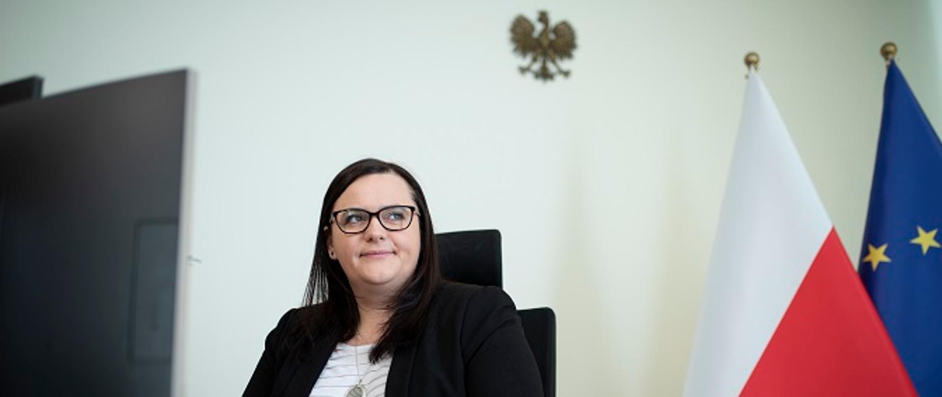 Minister Małgorzata Jarosińska-Jedynak przy biurku, w tle flagi Polski i UE.