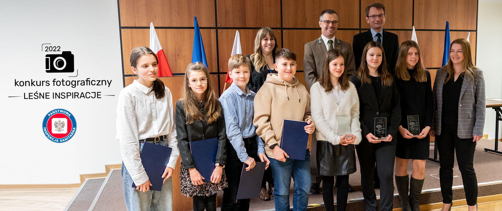 Grupa dzieci i dorosłych stają w dwóch rzędach. Dzieci, laureaci konkursu trzymają w rękach dyplomy i szklane statuetki.