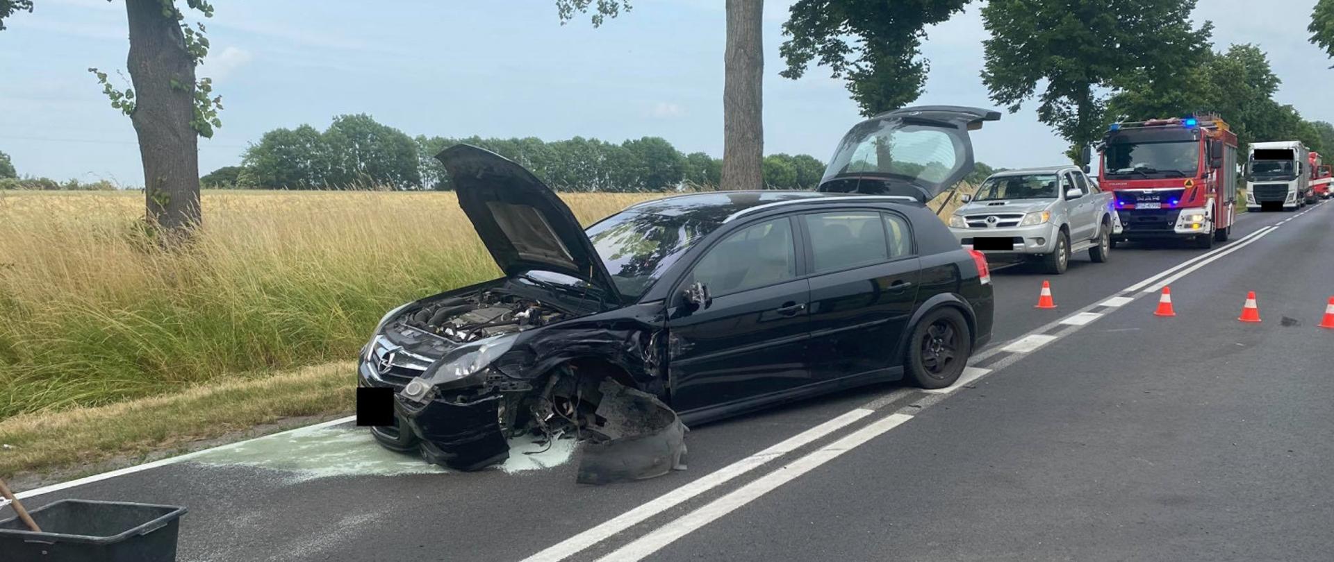 Widać uszkodzony ciemny samochód od przodu z otwartą maską na środku drogi, za nim kilka pojazdów