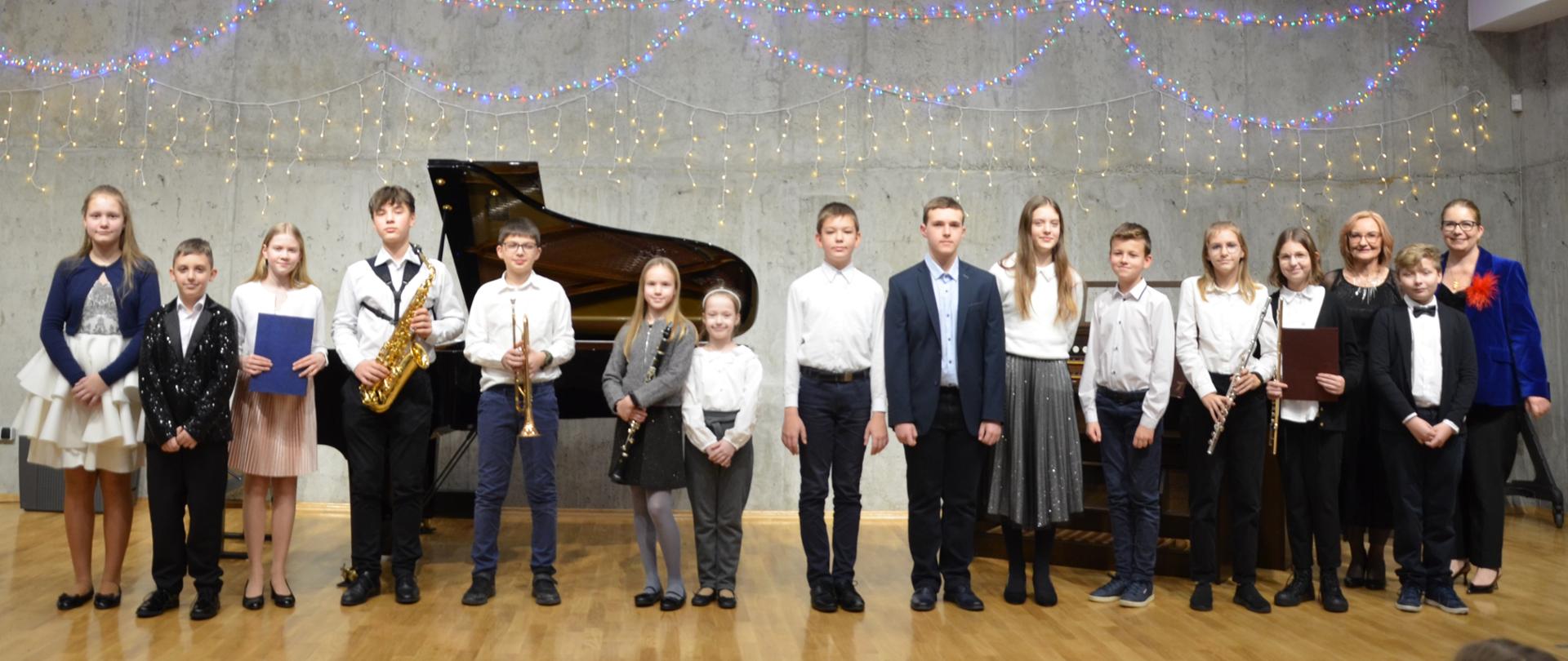Zdjęcie przedstawia grupę uczniów stojących na scenie sali koncertowej szkoły muzycznej