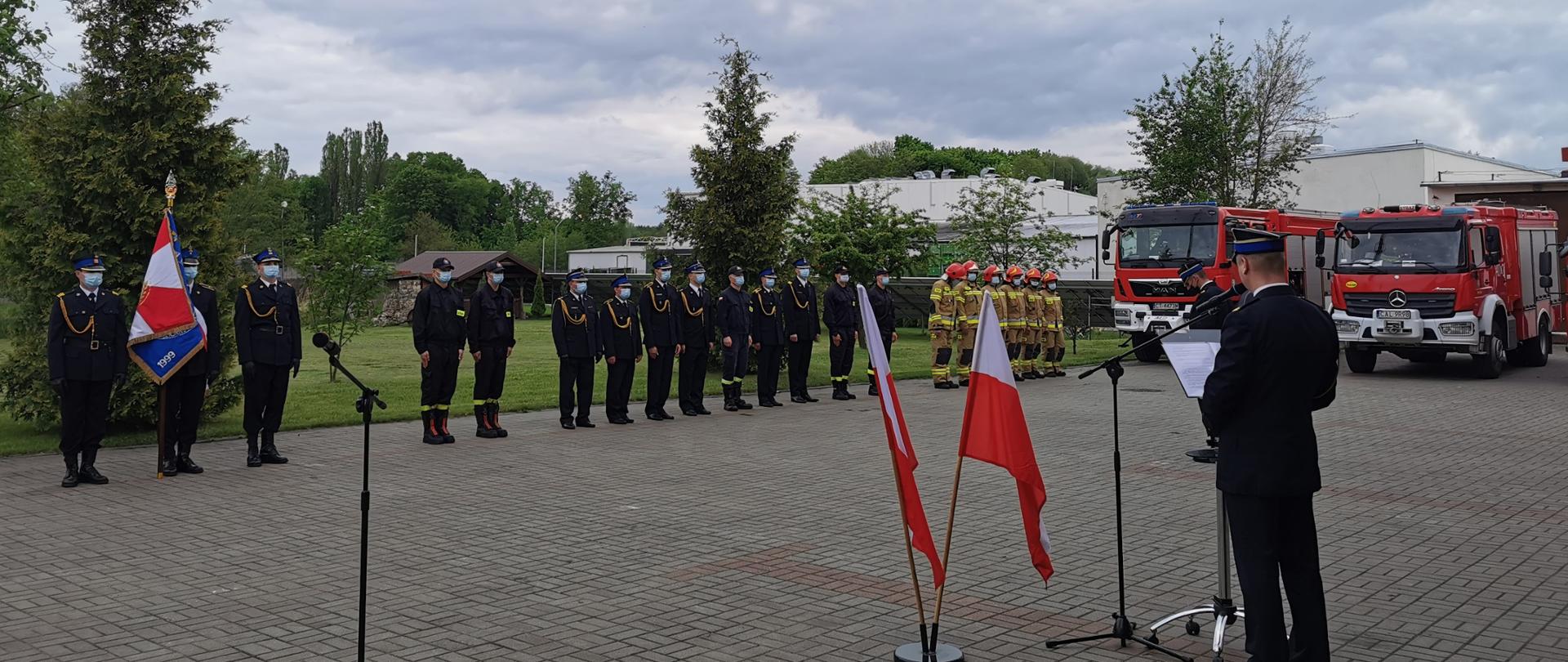 Zdjęcie przedstawia stojącego przy mównicy przemawiającego Komendanta Powiatowego PSP w Aleksandrowie Kujawskim. Na pierwszym planie stojak z dwiema flagami Polsk oraz przemawiający komendant. W tle stoją strażacy w szeregu. Po lewej stronie poczet sztandarowy. W prawej stronie zdjęcia znajdują się dwa wozy ratowniczo-gaśnicze.