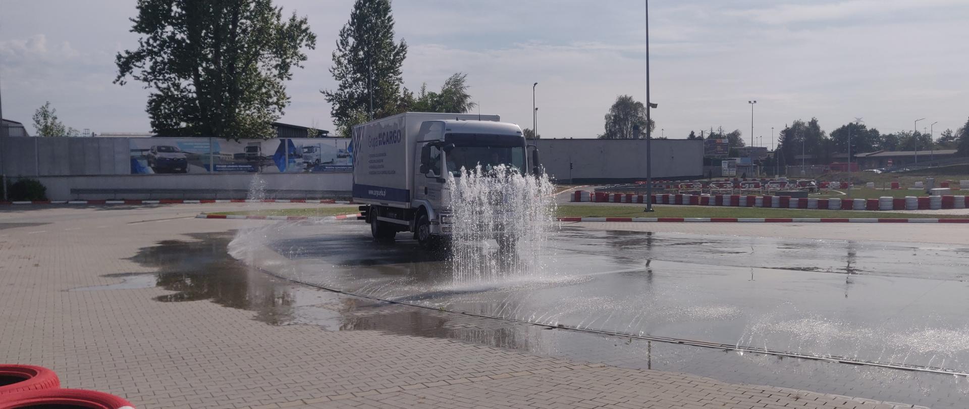 Po placu do nauki jazdy porusza się samochód ciężarowy. Przed nim i z boku ustawiona jest kurtyna wodna. Plac otoczony jest oponami ustawionymi w celu zabezpieczenia. W oddali widoczne budynki i drzewa.