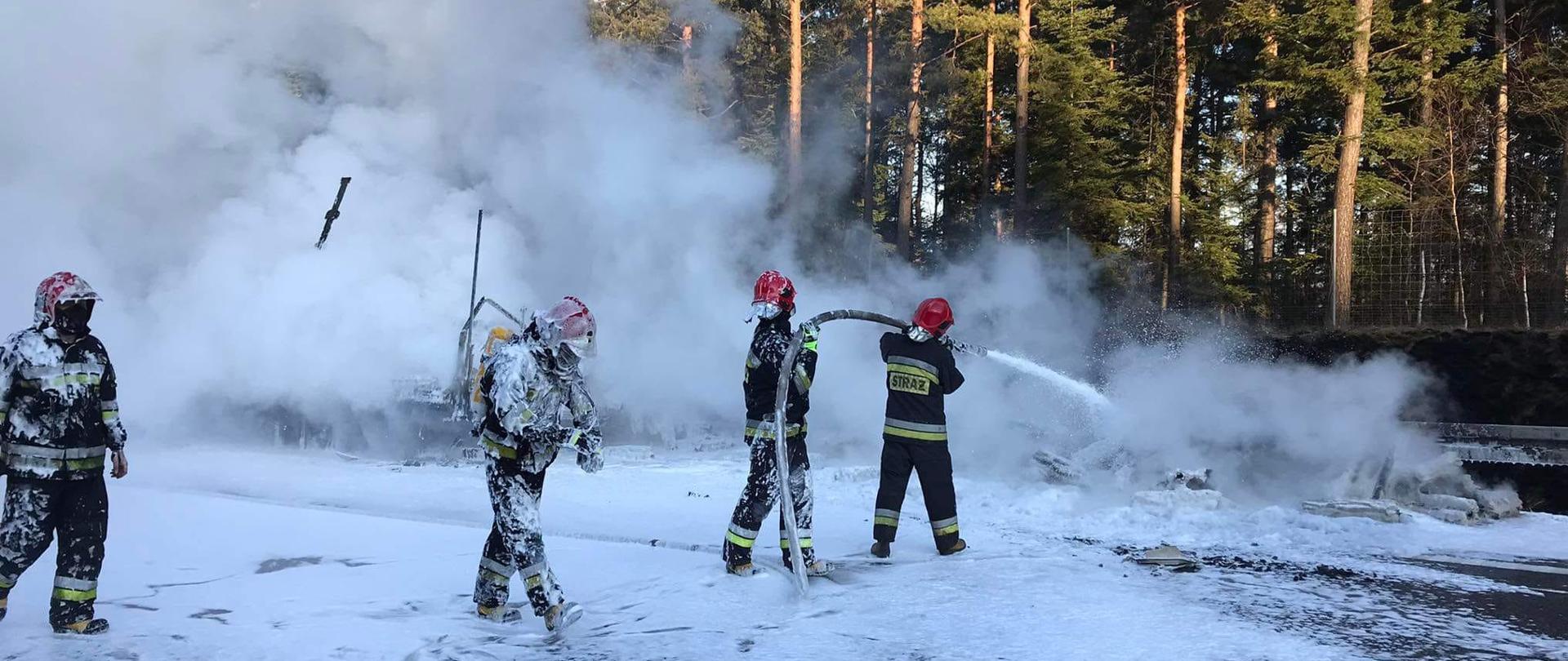 Strażacy pokryci pianą gaśniczą dogaszają nadpaloną folię. Na jezdni widać pianę gaśniczą, a w tle rosnące sosny.