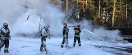 Strażacy pokryci pianą gaśniczą dogaszają nadpaloną folię. Na jezdni widać pianę gaśniczą, a w tle rosnące sosny.