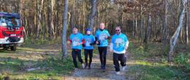 czterech mężczyzn w niebieskich koszulkach biegnie leśną ścieżką na ich twarzach widać uśmiechy po lewej stronie znajduje się czerwony samochód strażacki
