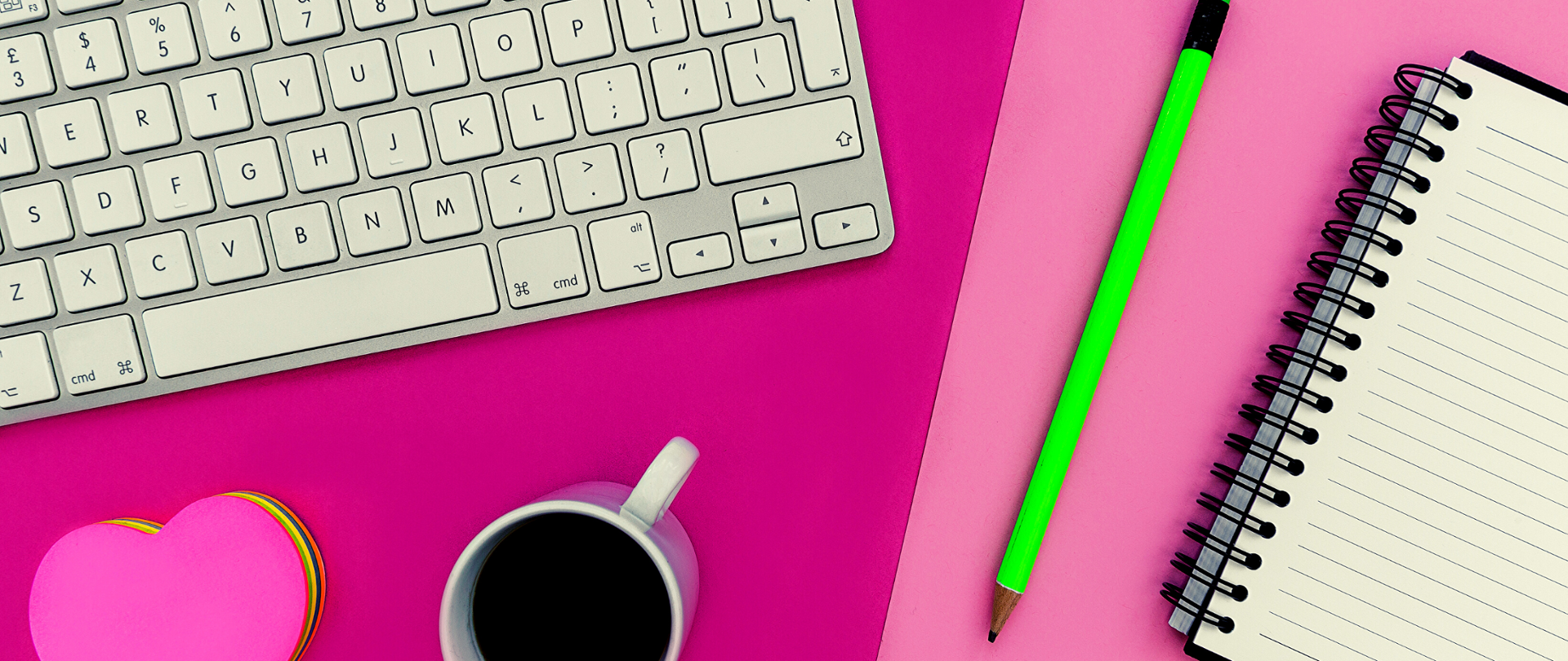 Na biurku leżą: klawiatura komputera, kubek z herbatą, ołówek i notes.