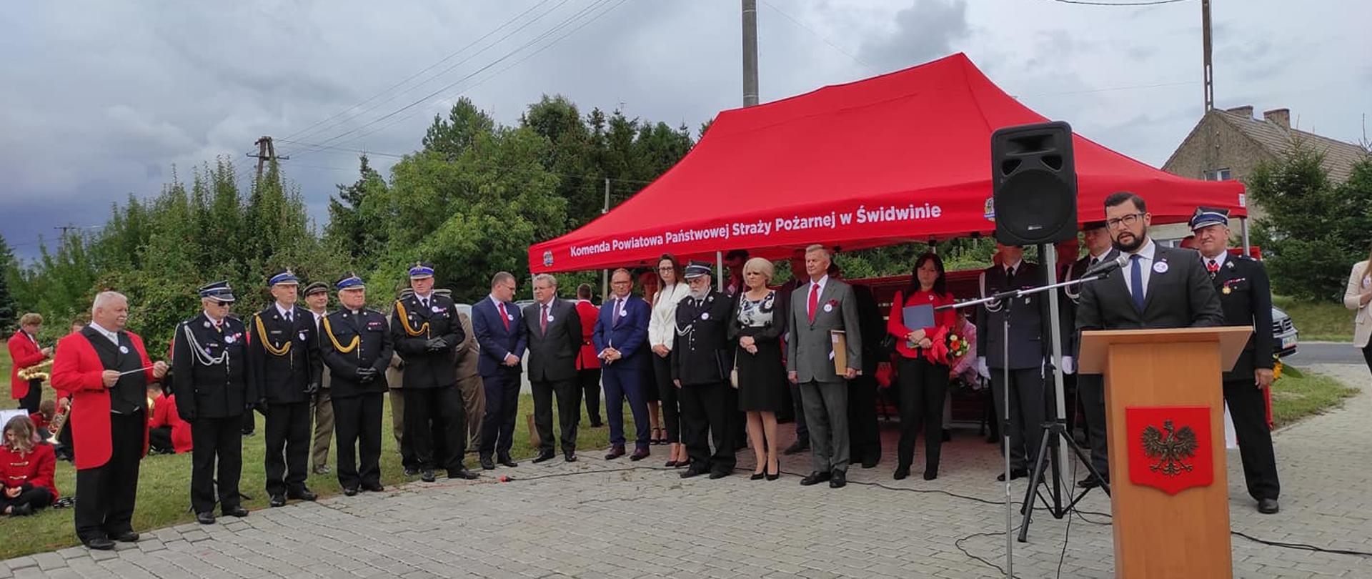 Zdjęcie przedstawia uroczysty apel z okazji 75-lecia jednostki OSP Rusinowo, strażacy są ubrani w mundury galowe. Przed mównica przemawia wicewojewoda.