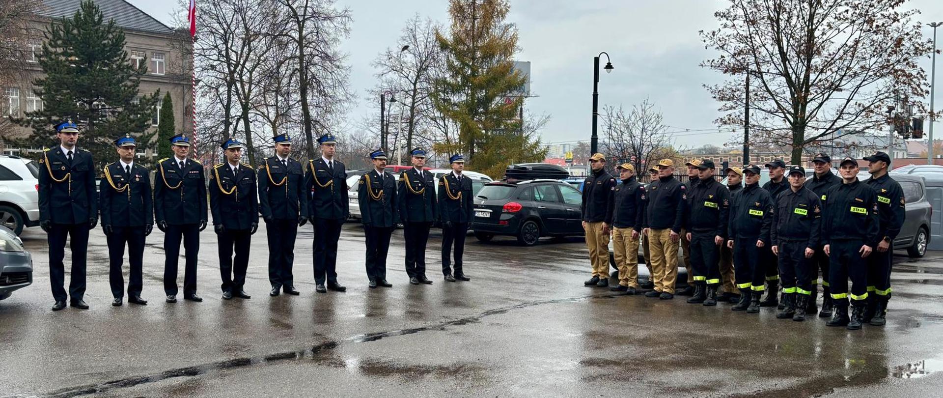 Na zdjęciu widoczne dwa pododziały strazaków. Centralnie w szeregu ustawieni strażacy w umundurowaniu galowym. Z ich lewej strony prostopadle ustawiony jest pododział w dwuszeregu w umundurowaniu koszarowym. Pogoda deszczowa. Strażacy ustawieni na placu komendy. W tle samochody osobowe. 