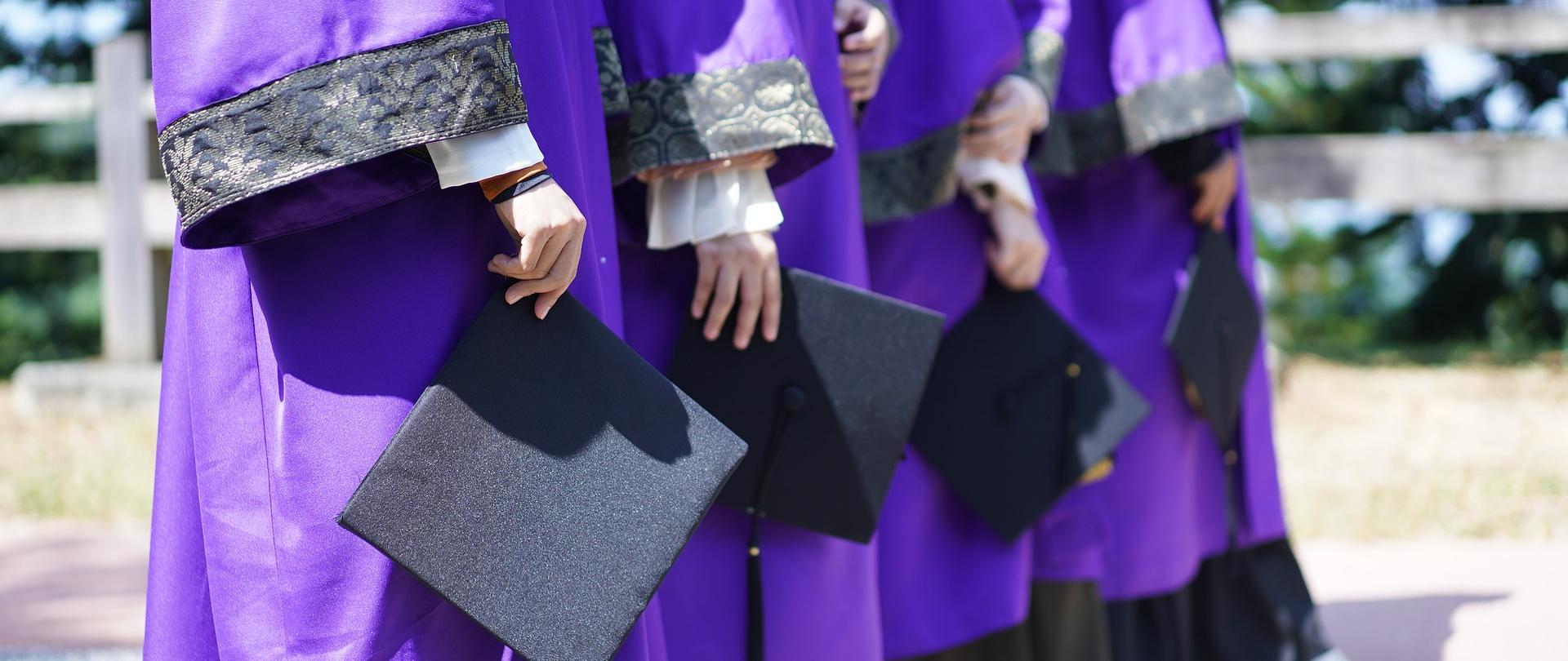 sylwetki 4 osób od pasa w dół w studenckich togach , trzymające w dłoniach birety