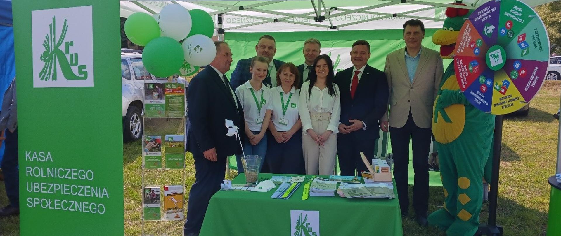 Grupa osób przedstawicieli instytucji i pracowników KRUS – ubranych elegancko lub w koszulki z identyfikatorami – pozuje do zdjęcia stojąc pod zielono-białym namiotem promocyjnym KRUS. Tło stanowią banery i stoisko informacyjne z materiałami promocyjnymi, broszurami i gadżetami. Namiot ozdobiony jest balonami i logotypem KRUS. Po prawej stronie znajduje się kolorowe koło fortuny oraz postać maskotki smoka w zielonym stroju, co dodaje wydarzeniu elementu zabawy i edukacji dla młodszych uczestników. 