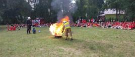Obóz MDP Brenno. Na plaży nad Jeziorem Białym trwają zajęcia z zakresu gaszenia pożarów przy pomocy gaśnic. Na środku ustawiona jest w wannaz palącą się cieczą. Występuje bardzo mocny płomień. Strazak w ubraiu specjalnym gasi pożar, używając do tego celu gaśnicy. W tle obozowicze przyglądający się tej akcji.