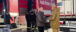 Pomorski komendant wojewódzki Państwowej Straży Pożarnej stoi z wojewodą pomorskim obok tirów do których ładowane są palety ze sprzętem pożarniczym.