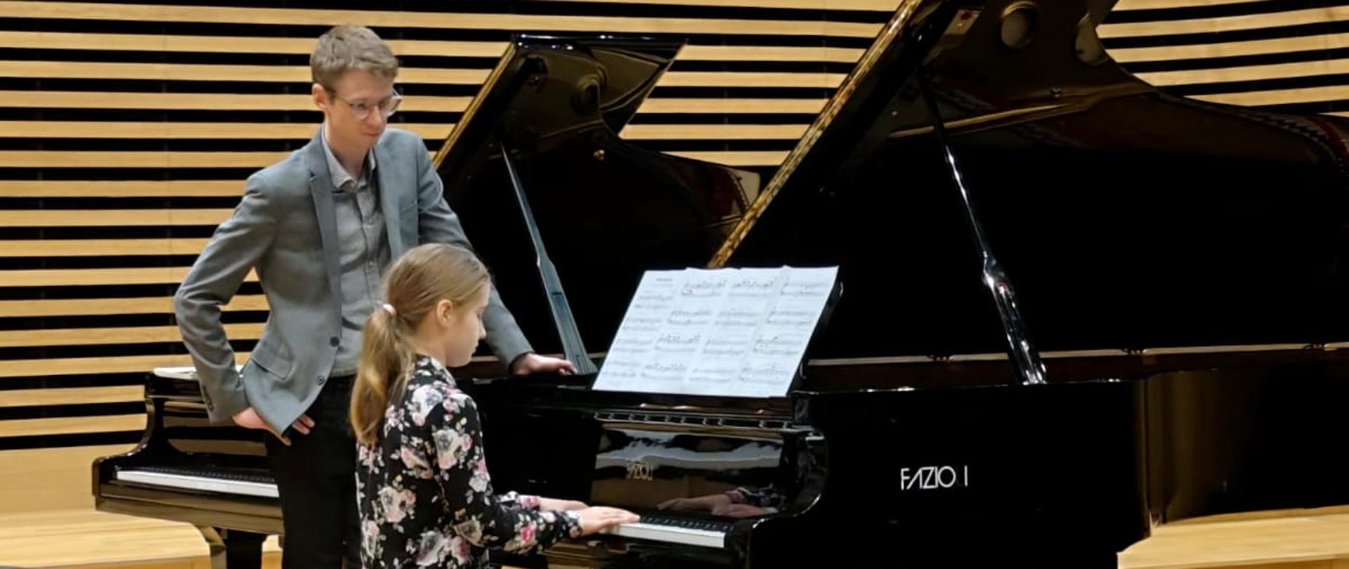 Dziewczynka gra na fortepianie, obok stoi mężczyzna.