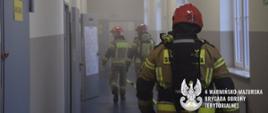 Strażacy w zadymionym korytarzu przeszukują pomieszczenia szukając poszkodowanych