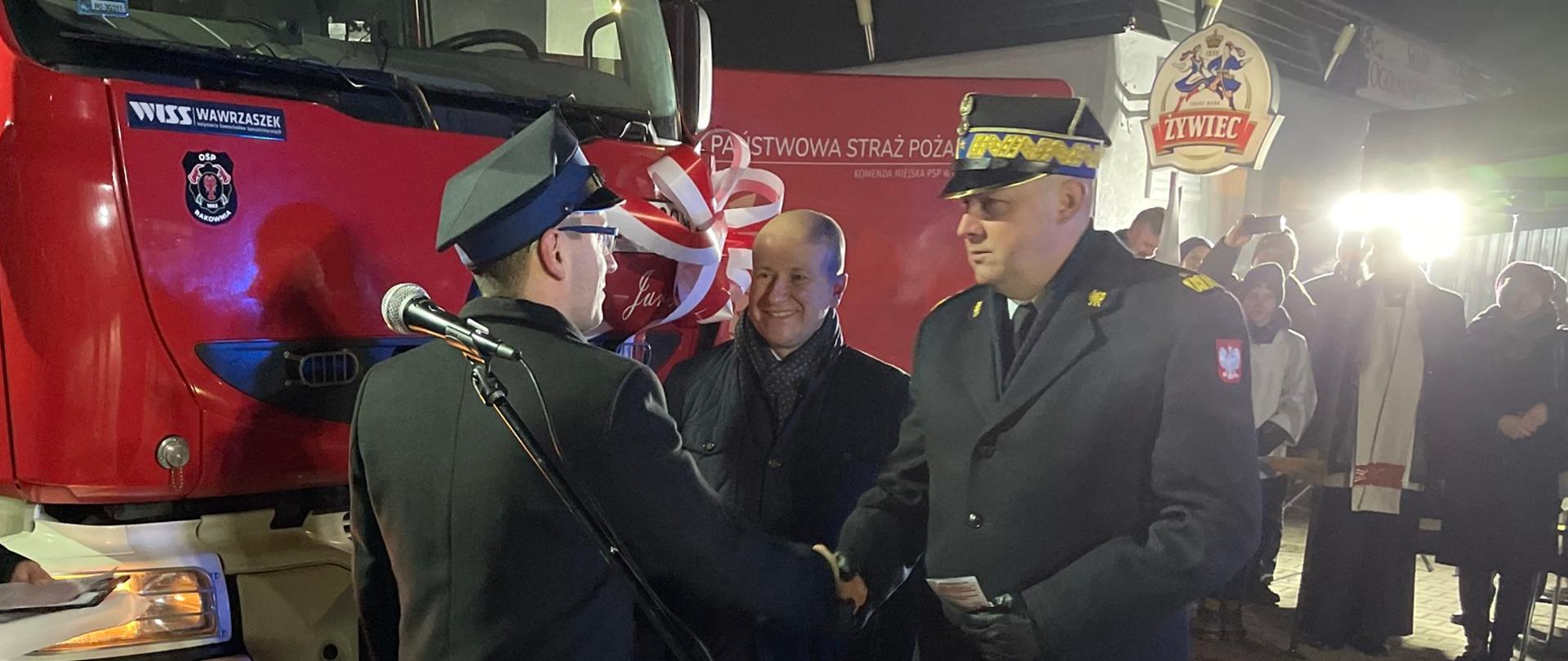 strażak w płaszczu i w rogatywce ściska dłoń drugiemu strażakowi, w tle czerwone samochody strażackie