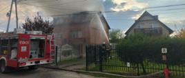 Budynek mieszkalny gdzie z poddasza unoszą się kłęby dymu obok strażacy prowadzący działania gaśnicze.