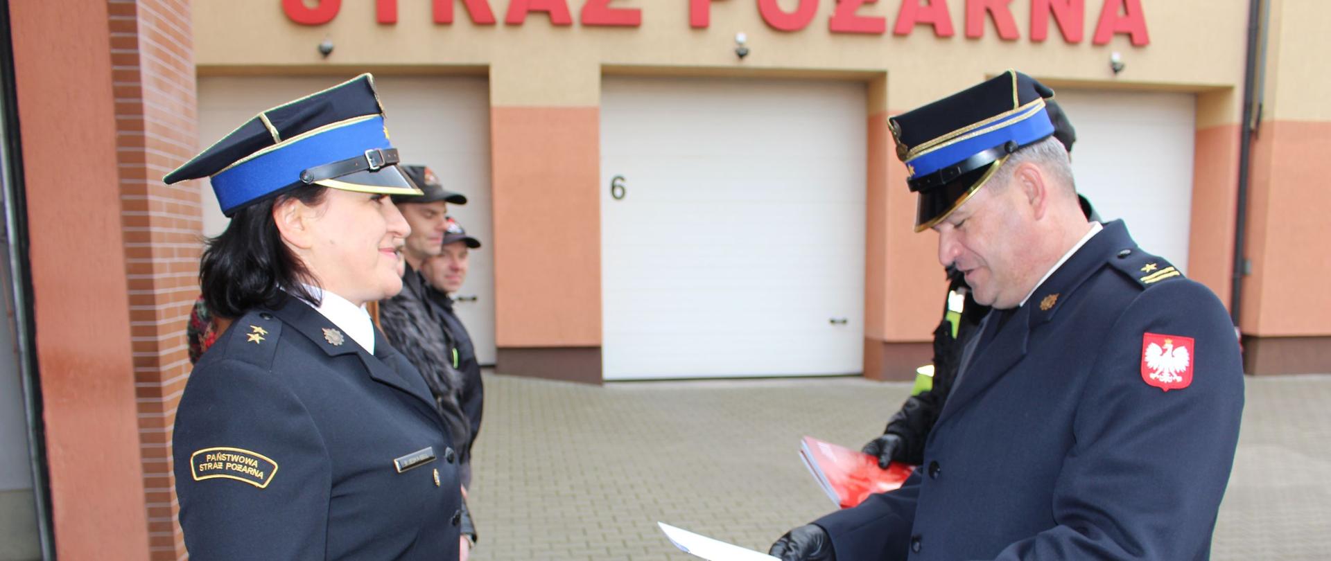 Zdjęcie przedstawia strażaków podczas uroczystej zmiany służby.
W tle budynki.
