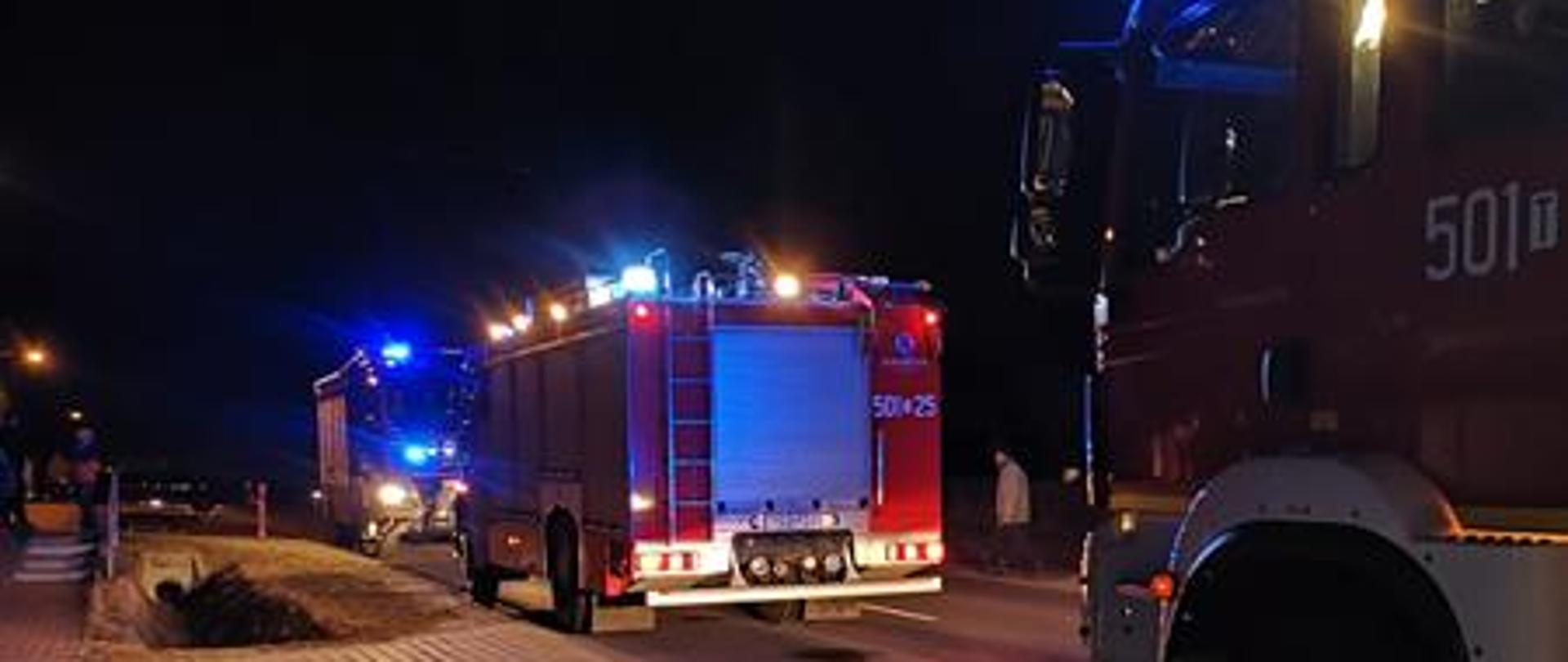 Na drodze wojewódzkiej obok posesji na której wystąpił pożar ustawione są pojazdy pożarnicze. Pojazdy maja włączoną sygnalizację świetlną.