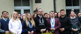 Przed budynkiem z żółtymi ścianami stoi grupa młodych ludzi, pośrodku minister Kulasek w czarnej bluzie z kapturem z napisem Parlament Studentów Rzeczypospolitej Polskiej, przed nimi zielone krzewy.
