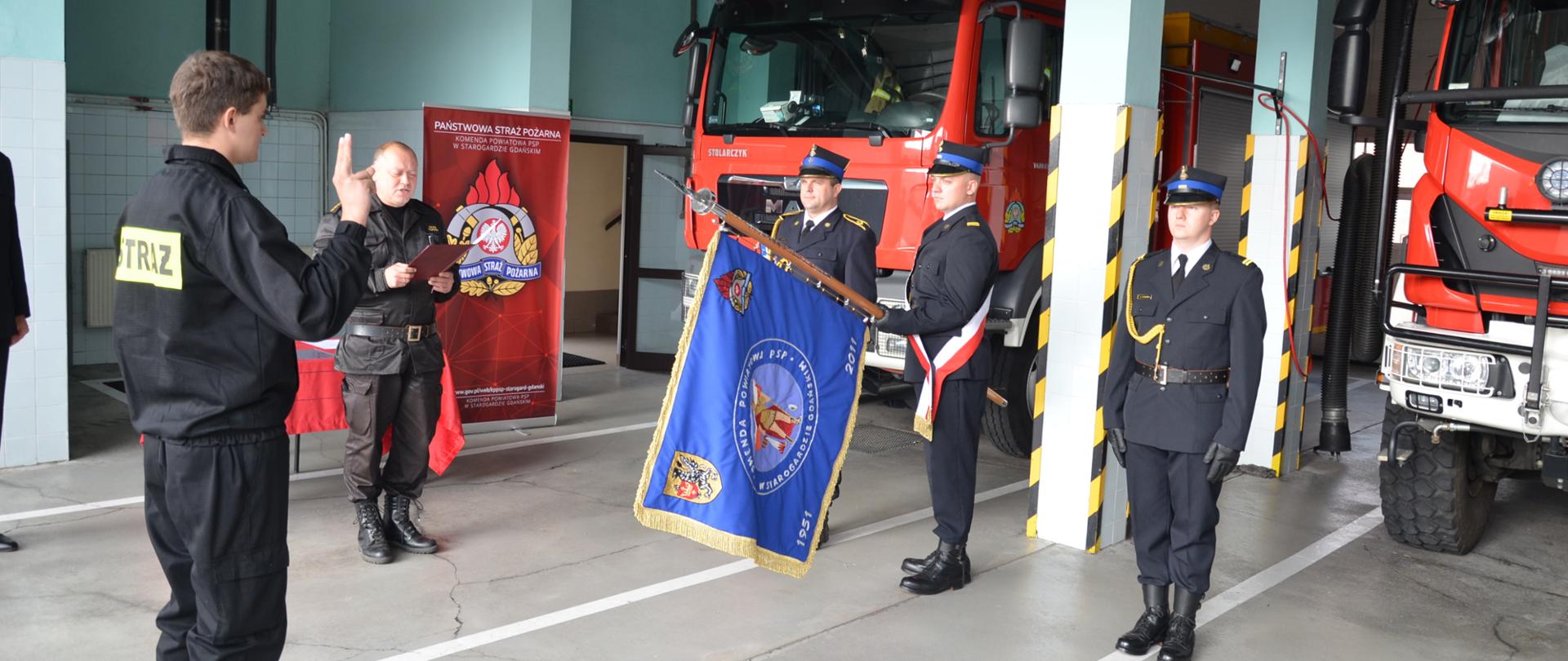 Zdjęcie przedstawia moment ślubowania, w tle samochody pożarnicze, poczet sztandarowy, strażak mówi rotę ślubowania, dowódca jednostki czyta rotę ślubowania