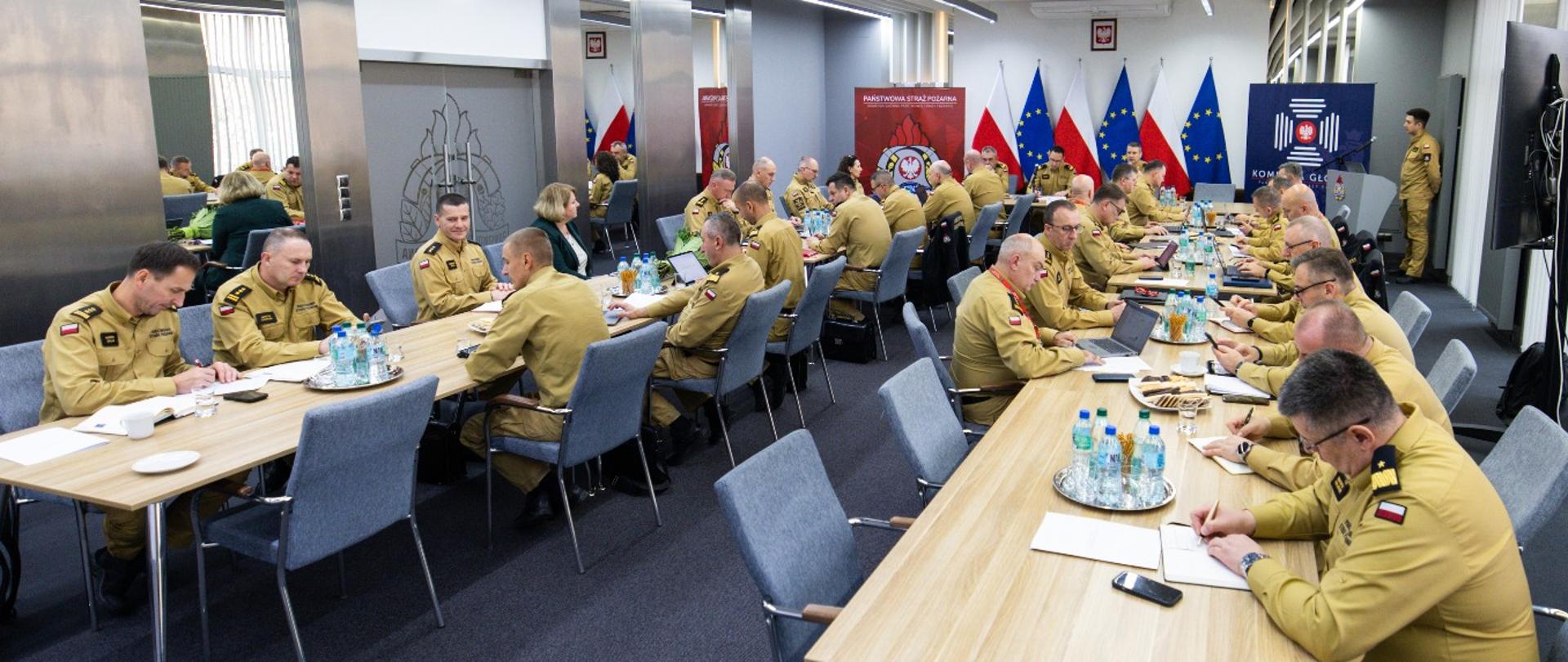Sala konferencyjna z kilkudziesięcioma strażakami PSP w żółtych mundurach służbowych siedzącymi przy długich stołach. W tle flagi narodowe i logo Państwowej Straży Pożarnej.