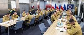 Sala konferencyjna z kilkudziesięcioma strażakami PSP w żółtych mundurach służbowych siedzącymi przy długich stołach. W tle flagi narodowe i logo Państwowej Straży Pożarnej.