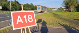 W tle dwupasmowa droga, na pierwszym planie czerwona tabliczka z napisem A18.