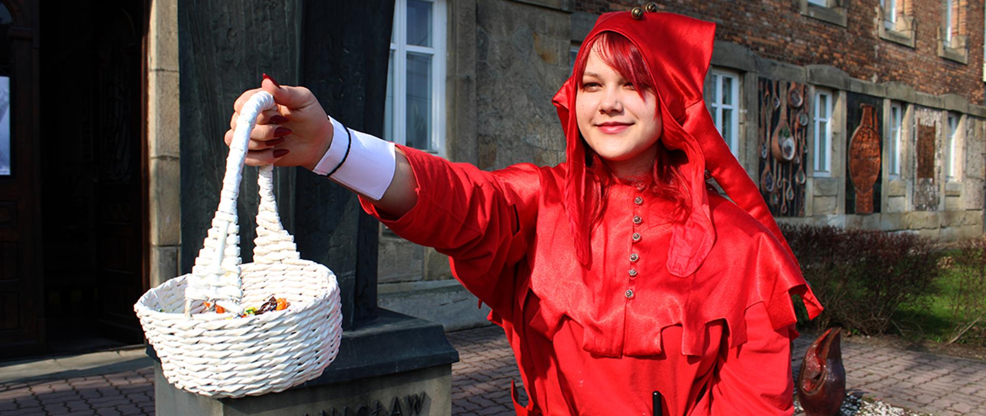 Fotografia została wykonana na zewnątrz, w przestrzeni miejskiej, przed budynkiem z ceglaną elewacją i oknami.
Na pierwszym planie widoczna jest osoba ubrana w czerwony kostium przypominający strój Stańczyka.
Osoba ta trzyma biały wiklinowy koszyk z cukierkami, wyciągnięty w kierunku obiektywu co jest symbolicznym zaproszeniem.