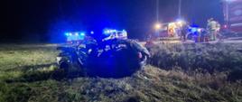 Na zdjęciu widoczny jest zniszczony pojazd osobowy, który podczas zdarzenia wypadł z drogi, pojazdy policji ZRM oraz straży pożarnje. Pomiędzy pojazdami widoczna jest torba medyczna oraz strażacy.