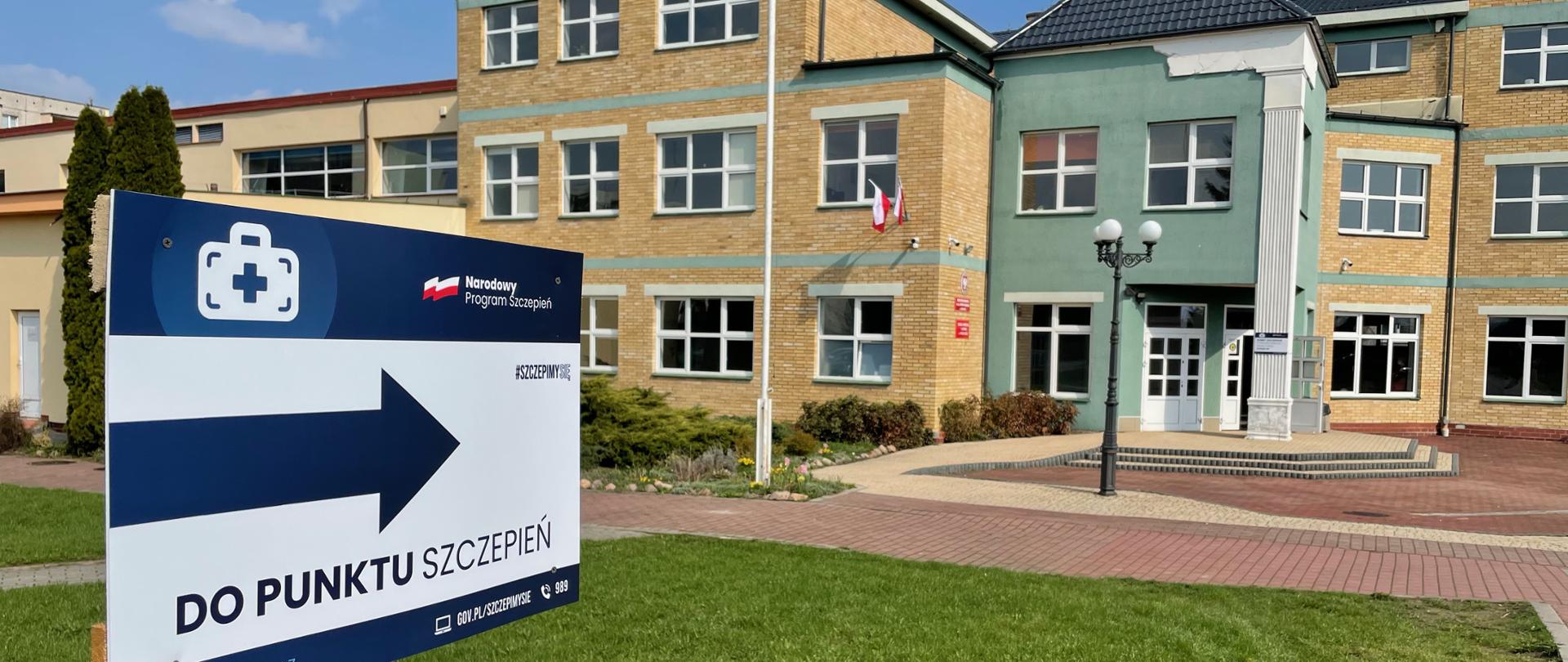 Zdjęcie tablicy kierującej do punktu szczepień. W tle budynek zespołu szkół powiatowych w Przasnyszu.