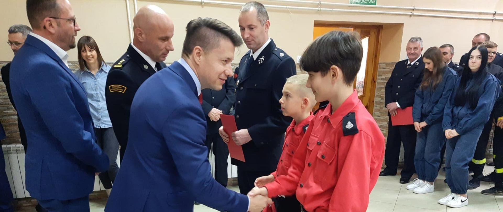 Zdjęcie przedstawia posła na Sejm RP Bartłomieja Dorywalskiego, który wręcza promesy dla Młodzieżowych Drużyn Pożarniczych. Posła asystują zastępca świętokrzyskiego komendanta wojewódzkiego PSP st. bryg. Grzegorz Rajca, starosta jędrzejowski Paweł Faryna.